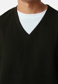 Tmavě zelený svetr s výstřihem do V vyrobený z texturované látky. Má žebrovaný límec a vrstvené bílé tričko viditelné u výstřihu.