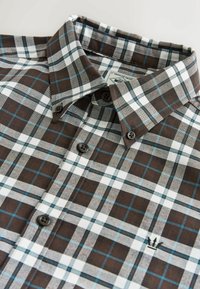 Chemise à carreaux marron et blanche avec des accents bleus, col boutonné et boutons foncés. Dispose d'un petit logo brodé au-dessus de la poche.