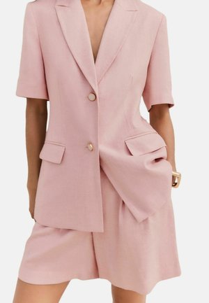 Nő világos rózsaszín, testre szabott blézerben arany gombokkal és hozzáillő, magas derekú rövidnadrágban, kezét a zsebében tartja, fehér háttér előtt.