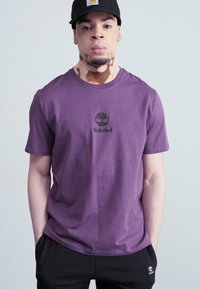 Violet medvilninė marškinė su apvaliu kaklu, ant krūtinės centre yra juodas Timberland logotipas. Dėvimi su juodomis kelnėmis.