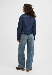 Tamsiai mėlyna džinsinė striukė su formos liemeniu, derinama su šviesiai mėlynomis plačiomis kelnėmis. Pabrėžia du matomi užpakaliniai kišeniai ir priekiniai mygtukų užsegimai.