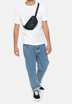 Mand iført hvid t-shirt, blå jeans, sort skuldertaske og sorte sneakers går mod ensfarvet baggrund.