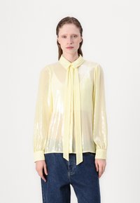 Transparante gele blouse met pailletten, lange mouwen, kraag en strik. Zachte textuur, losse pasvorm, gedragen met donkerblauwe jeans.
