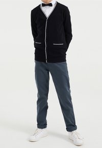 Černý pletený cardigan s bílými detaily, zapínání na knoflíky a kapsami, ve spojení se šedými kalhotami a bílými teniskami. Pod ním je oblečený motýlek a košile s límečkem.