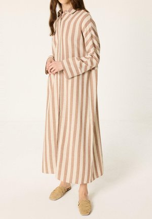 Femme portant une longue robe rayée verticale beige et blanche avec des manches longues et des chaussures beige tissées sans lacets, debout devant un fond uni.