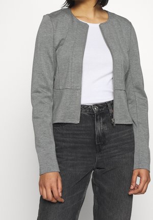 Persona con una chaqueta gris con cremallera sobre una camiseta blanca combinada con unos vaqueros negros de cintura alta contra un fondo liso.