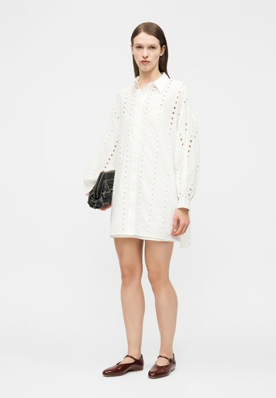 Kvinde iført en hvid langærmet kjole med hulmønster, der holder en sort håndtaske og har mørkebrune Mary Jane-sko på, stående på en hvid baggrund.