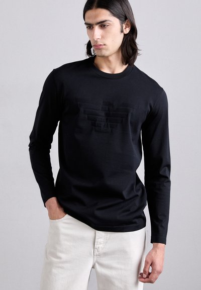 Melna garām piedurknēm T-kreklu no gludas auduma, ar uzspiestu logotipu priekšpusē, apvienots ar gaišām biksēm.