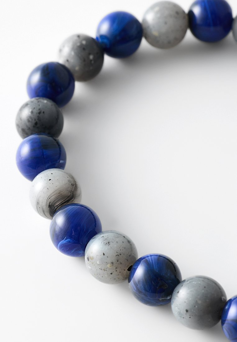Bracciale di perle rotonde che alternano pietre levigate di un profondo blu e pietre grigie opache macchiate su uno sfondo bianco.