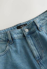 Jean en denim bleu clair avec un bouton en métal portant l'inscription « NEXT DENIM SINCE 1982 » et une étiquette en tissu noir avec « NEXT » à l'intérieur de la ceinture.