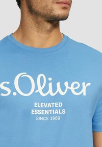 Light blue cotton t-shirt featuring a round neckline and short sleeves. The front displays "s.Oliver" and "ELEVATED ESSENTIALS SINCE 1969" in white.