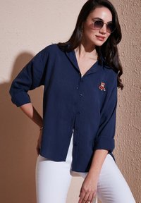 Navy blue button-up shirt made of lightweight fabric, featuring a small embroidered bear design on the chest, with rolled sleeves.
