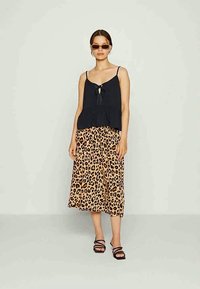 Mulher vestindo uma blusa preta sem mangas, saia midi de estampa de leopardo, sandálias pretas de tiras e óculos de sol, de pé contra um fundo claro liso.