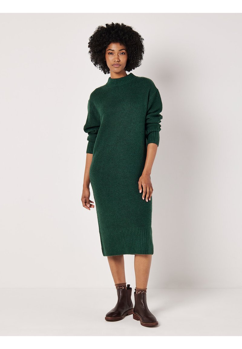 Grünes Strickkleid mit hohem Kragen, lockeren Ärmeln, geripptem Saum und gerader Silhouette; kombiniert mit braunen Stiefeletten.