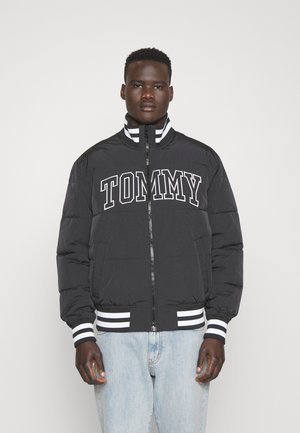 Chaqueta acolchada negra con letras "TOMMY" en el pecho, cuello y puños a rayas blancas, y cierre de cremallera. Combinada con jeans de denim claros.