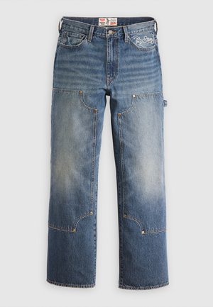 Blue denim jeans with a loose fit, featuring large patched pockets, brass hardware, and distinct stitching patterns. Embroidered logo on waistband.