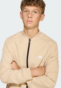 Béžová žíňovaná bomber bunda s čiernym zipsom, s bielym logom Adidas na ľavej hrudi. Elastické manžety a okrúhly výstrih.