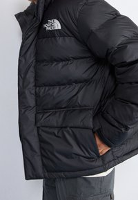 Giubbotto in piuma nero con design trapuntato, dotato di tasca frontale con patta e logo The North Face sul petto. Tessuto con texture, isolamento caldo.