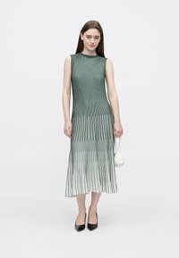 Vestido verde plissado com riscas verticais, design sem mangas, silhueta fluida, combinado com uma pequena mala branca e saltos pretos pontiagudos.