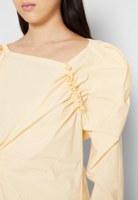 Kvinna som bär bländande gulblus med rynkade detaljer och puffärmar, visad från halsen till mitten av överkroppen mot en enkel bakgrund.