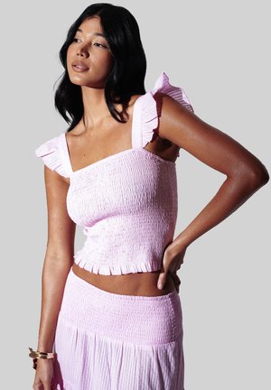 Vrouw met halflang zwart haar, gekleed in een lichtroze smock cropped top en bijpassende rok, poseert met één hand op haar heup.