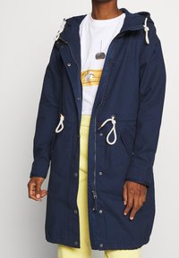 Mörkblå parka med huva, utrustad med dragsko, framslutning med dragkedja och fickor. Materialet verkar slätt med en avslappnad design.