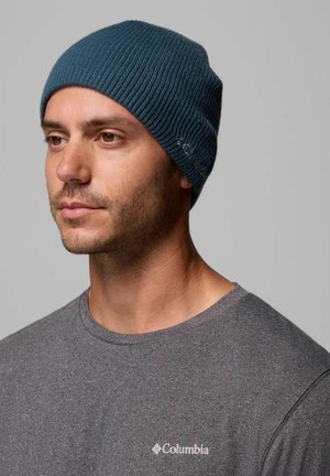 Uomo con barba corta che indossa un berretto di lana blu e una maglietta grigia Columbia, guarda leggermente verso sinistra su uno sfondo grigio uniforme.