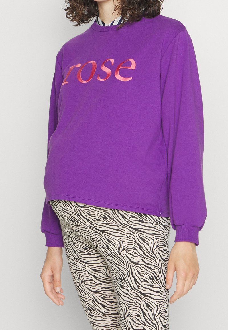 Personne portant un sweat-shirt violet avec le texte "rose" et un pantalon imprimé zèbre noir et blanc, debout sur un fond uni.