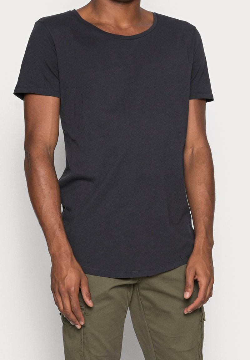 Homme portant un t-shirt noir uni à manches courtes et un pantalon vert olive, debout devant un fond blanc.