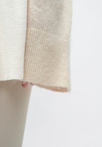 Beežist kudumiga kampsun soonikkoes tekstuuriga, millel on pehme, karvane ääris ja kerge värvimuutus varrukate otstes, sobitatud õhukeste hallide pükstega.