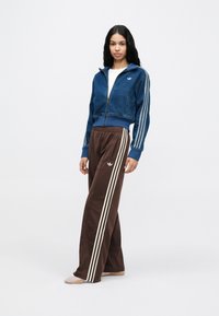 Jachetă scurtă albastră cu fermoar, textură de denim și dungi albe, asortată cu pantaloni maro cu croială largă și dungi albe pe lateral.