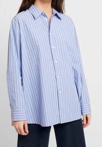 Světle modrá košile na knoflíky s tenkými bílými pruhy, kapsou na levé straně, dlouhými rukávy se pruhovanými manžetami, vyrobená z měkké tkaniny.