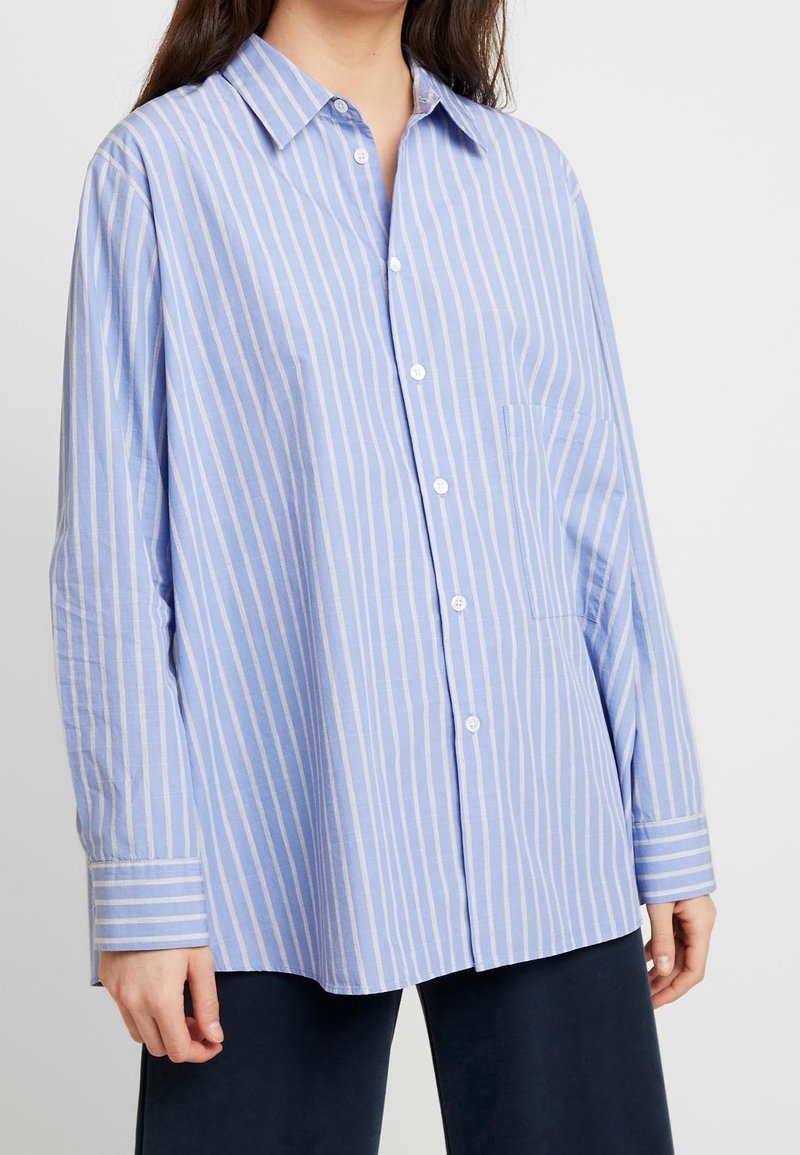 Světle modrá košile na knoflíky s tenkými bílými pruhy, kapsou na levé straně, dlouhými rukávy se pruhovanými manžetami, vyroben á z měkké tkaniny.