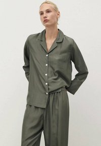 Olivengrünes Seiden-Pyjama-Set mit einem knöpfbaren Hemd mit Tasche und langen Ärmeln, kombiniert mit Hosen mit elastischem Bund. Weicher Stoff.