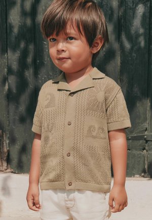 Copil mic cu păr scurt castaniu, purtând o cămașă bej cu mâneci scurte și model și pantaloni albi, stând în fața unei uși de lemn verde închis.