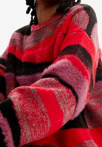 Pull rayé rouge et noir avec des textures variées, incluant des accents duveteux. Présente des rayures horizontales dans des tons de rouge, rose et gris.