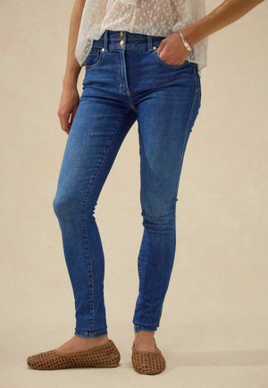 Kvinde iført blå højtaljede skinny jeans, en gennemsigtig hvid blonder top, brune flettede fladsko og et sølvarmbånd med hånden i lommen.