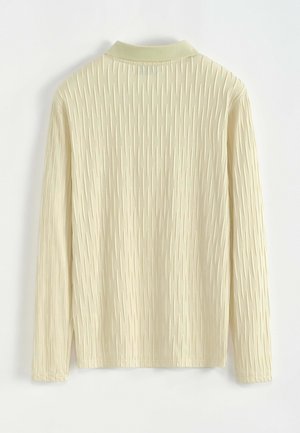 Haut à manches longues jaune pâle avec un motif texturé de lignes verticales, doté d'un col à revers et de poignets légèrement élastiqués.