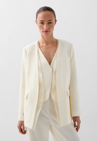 Blazer crema a medida con una textura suave, de frente abierto, con dos bolsillos y un corte relajado, superpuesto sobre una blusa crema a juego.