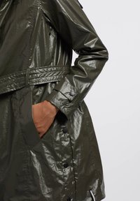 Olijfgroene, glanzende regenjas met een ceintuur, drukknoopsluiting en een zak. De stof heeft een gekreukt textuur met een glanzende afwerking.