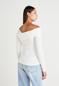 Witte langemouwen top met een off-the-shoulder ontwerp. Gladde stof, aansluitende pasvorm en een effen oppervlakte. Combinatie met lichtblauwe jeans.