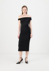 Svart off-shoulder kjole med tettsittende silhuett, kne-lengde hem og rynket tekstur. Båret med svarte spisse hæler, med ankelremmer.