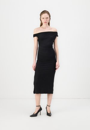 Svart off-shoulder kjole med tettsittende silhuett, kne-lengde hem og rynket tekstur. Båret med svarte spisse hæler, med ankelremmer.