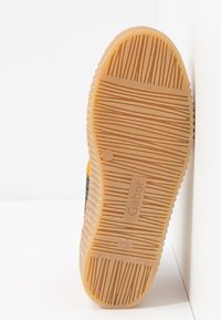 Semelle d'une chaussure Gabor jaune et noire avec une bande de caoutchouc texturée et rainurée pour l'adhérence, photographiée sur un fond blanc.