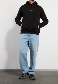 Hombre vestido con sudadera con capucha negra y logo, jeans de mezclilla azul claro y zapatillas negras, de pie con las manos en los bolsillos de la sudadera, contra un fondo blanco.