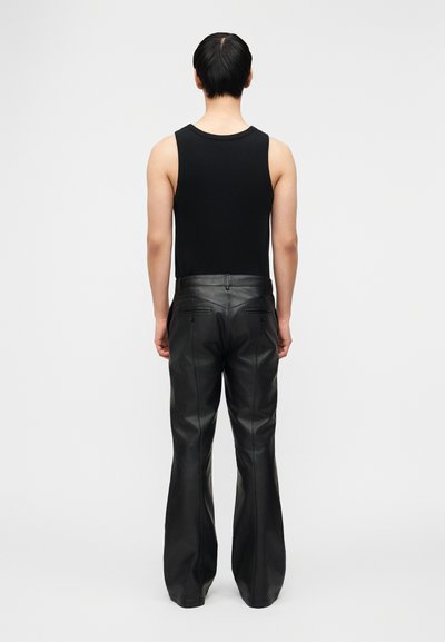 Άτομο με μαύρο αμάνικο τοπ και μαύρο δερμάτινο παντελόνι με φαρδιά καμπάνα, κοιτάζοντας προς τα πίσω, μπροστά σε απλό φόντο.