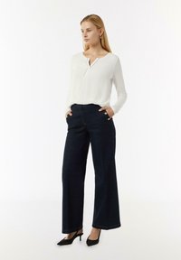Weite, dunkle Jeanshose mit goldenen Knopfakzenten, kombiniert mit einem weißen Langarmoberteil mit Henley-Ausschnitt. Schwarze Absätze runden den Look ab.