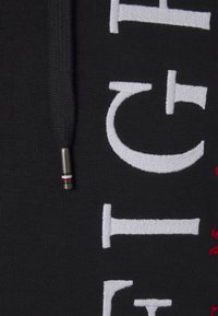 Felpa in tessuto nero con grande testo ricamato bianco e rosso; include un cappuccio nero con un laccio in metallo argento e accenti rossi.
