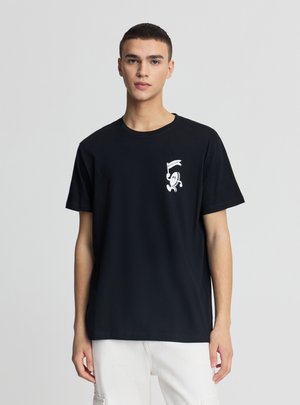 Giovane uomo in piedi, rivolto verso l'obiettivo, indossa una maglietta nera con una piccola grafica bianca di uno scheletro che tiene una bandiera sul petto e pantaloni bianchi.