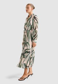 Femme debout de profil portant une robe midi beige avec des motifs de feuilles vertes, des manches longues, un devant boutonné et des sandales à talons noires.